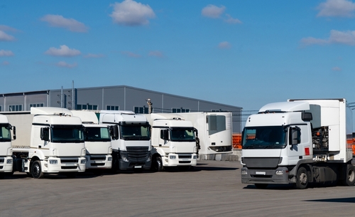 A row of parked white semi-trucks lined up in an industrial lot outside a warehouse under a blue sky.