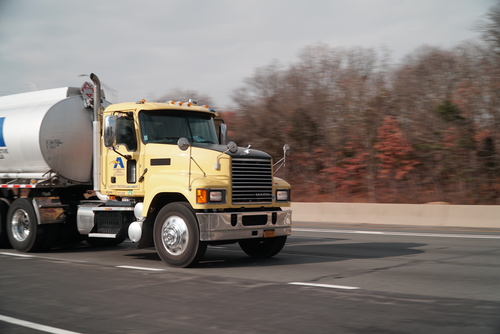 A yellow tanker truck driving at speed on a highway with blurred trees in the background.