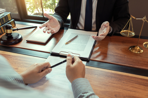 A lawyer meets with a client at a wooden desk, with legal documents, a pen, scales of justice, and a gavel placed on the table.