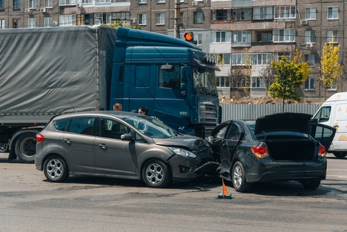 A truck accident scene showing two cars badly damaged in a collision, with a large blue semi-truck in the background and bystanders nearby.