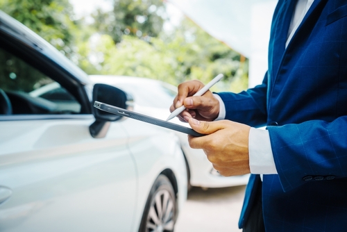 Insurance adjuster in a suit inspecting a damaged car and recording notes on a tablet during claim assessment.