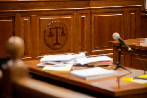 Close-up view of a courtroom desk with legal documents, microphone, and the scales of justice emblem carved into the wooden panel.