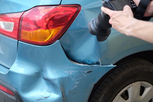 Close-up of a blue car’s rear corner with a deep dent and scratches while a person photographs the damage.