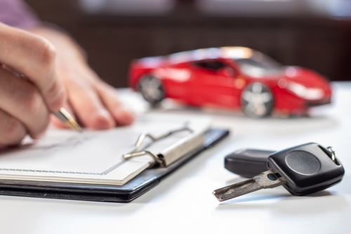 Close-up of a person signing car insurance paperwork with a key and a small red car model on the desk.