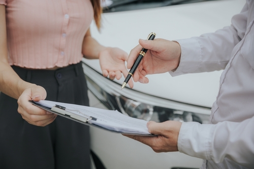 Two people standing by a white car, one holding a clipboard and pen while the other gestures with their hands.