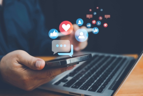 A person using a smartphone in front of a laptop, with colorful social media icons such as likes, hearts, comments, and followers floating above the phone screen, representing online engagement and digital communication