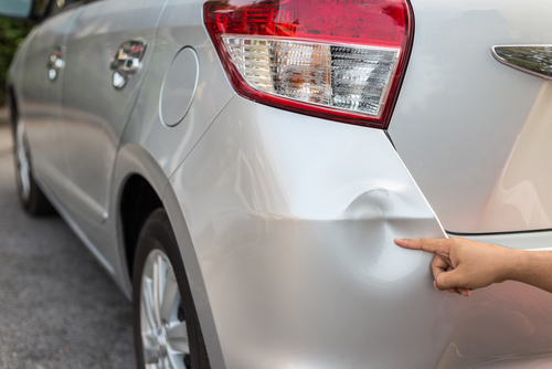 Hand pointing to a round dent on the rear door of a silver car, highlighting minor vehicle damage.